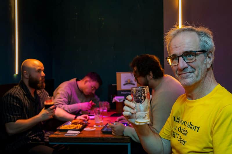 Ein Mann in einem gelben Hemd hält ein Glas in einem schwach beleuchteten Raum, in dem im Hintergrund Menschen an einem Tisch sitzen.