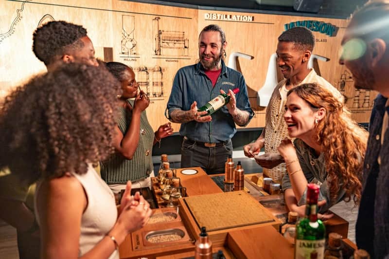 A group of people enjoying a whiskey-tasting session at a distillery, with a presenter holding a bottle of Jameson.