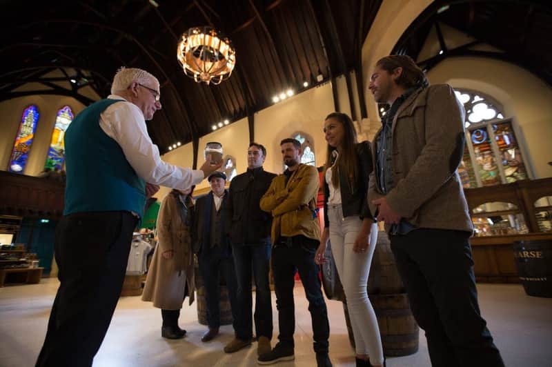 Pearse Lyons Distillery: Guided Tour with 3 Tastings A man in a teal vest explains something to a group of five people inside a building with wooden beams and barrels.