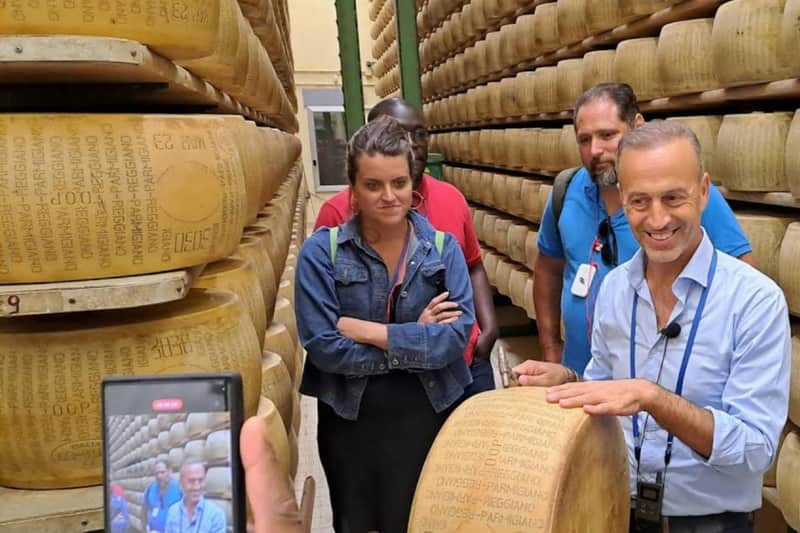 Noceto: Parmesan Cheese Factory Guided Tour A group of people stands in an aisle between shelves stacked with large wheels of cheese, one man smiling and holding a cheese wheel.