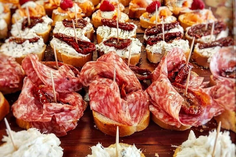Eine Nahaufnahme von verschiedenen Vorspeisen auf kleinen Brotscheiben, belegt mit Fleisch, Käse, getrockneten Tomaten und Garnierungen.