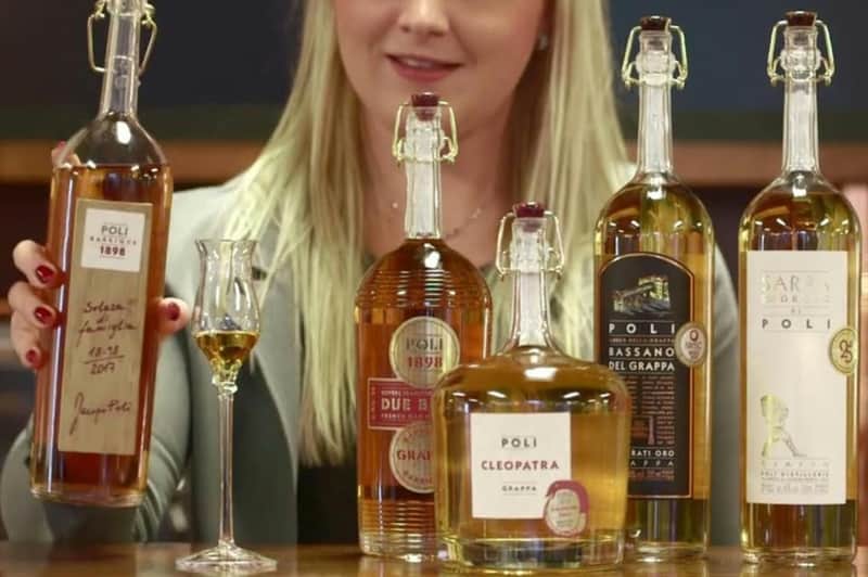 Eine Frau hält ein Glas Grappa hinter drei Flaschen Poli Grappa, darunter Poli Cleopatra, Due Barili und Bassano del Grappa.