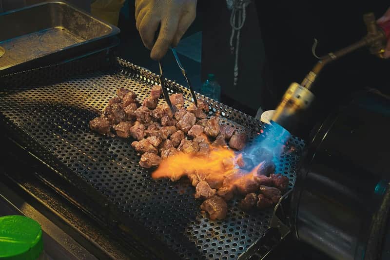 Kuromon Market, Sennichimae & Dotonbori: Guided Food Tour A person sears chunks of meat on a perforated grill using tongs and a blowtorch with a visible flame.