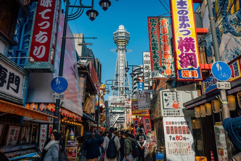 Osaka: Tennoji Ward & Shinsekai Guided Walking Tour Bustling street scene in Japan with people, colorful advertisements, shops, and a tall tower in the background under a clear blue sky.