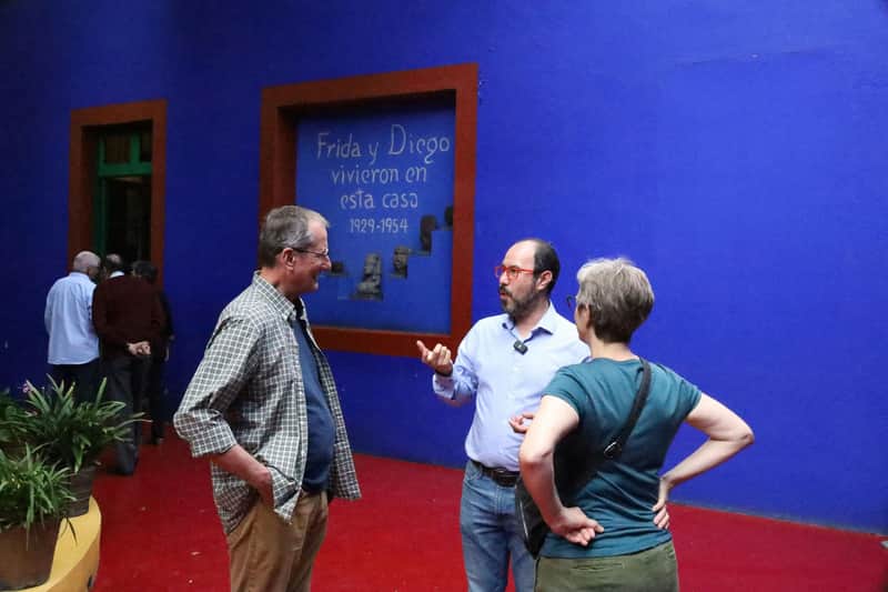 Three people conversing in front of a blue wall displaying text about Frida and Diego, with a red floor in the foreground.