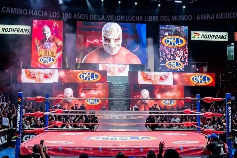 A wrestling ring surrounded by an audience, with large screens displaying a masked wrestler and CMLL logos.