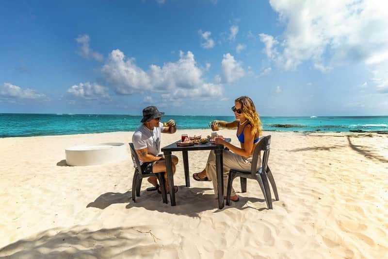 Two people sitting at a small table on a sandy beach, enjoying drinks and food under a clear blue sky with ocean in the background.