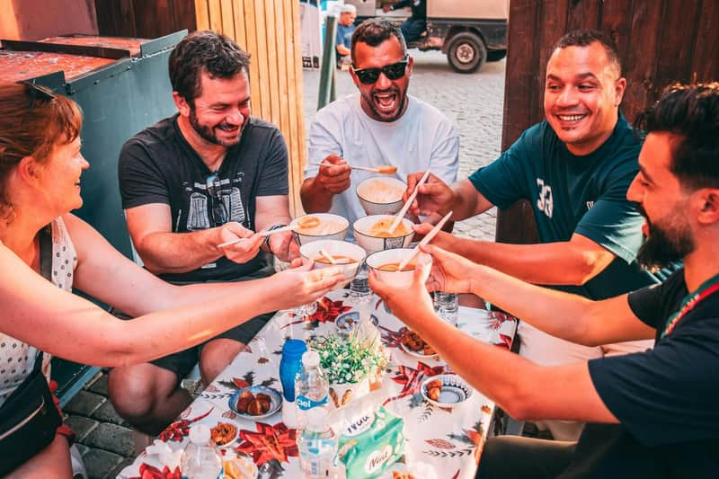 Marrakesh: Tour guidato serale del cibo di strada + degustazioni (Marrakech) Quattro uomini siedono a un tavolo all'aperto, sorridendo e condividendo ciotole di zuppa, con vari cibi e bevande sul tavolo.
