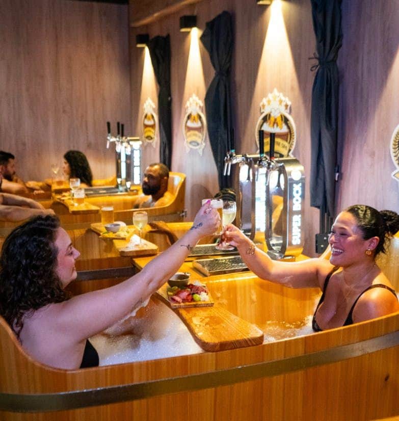 Beer & Wine Spa Lisbon: Entry + Drinks People in wooden hot tubs, toasting with drinks. Each tub has a wooden tray with food, beer taps, and a wall logo behind.