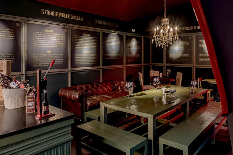 Beer Museum Lisbon: Guided Tour + Food & Craft Beer Tastings A cozy tasting room with a green table, benches, a leather couch, a chandelier, and informational panels on the walls.