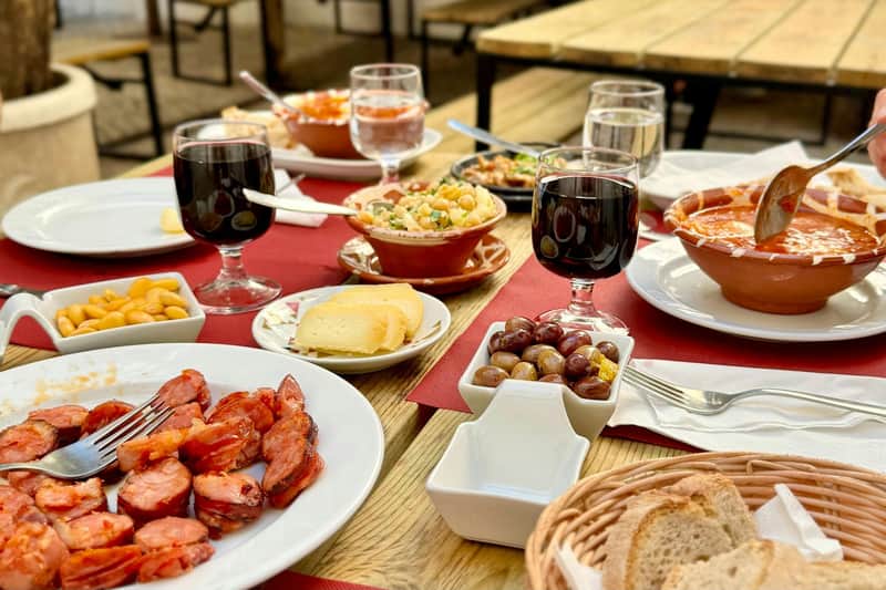 Lisbon: Foodie's Walking Tour A table set with various dishes, including sausages, cheese, olives, salad, and glasses of red wine, in an outdoor dining area.