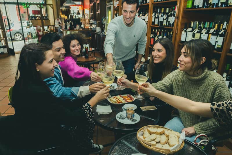 Porto: Guided Evening Food Tour Five people are clinking wine glasses around a table with food, in a cozy, dimly-lit wine bar with shelves of wine bottles.
