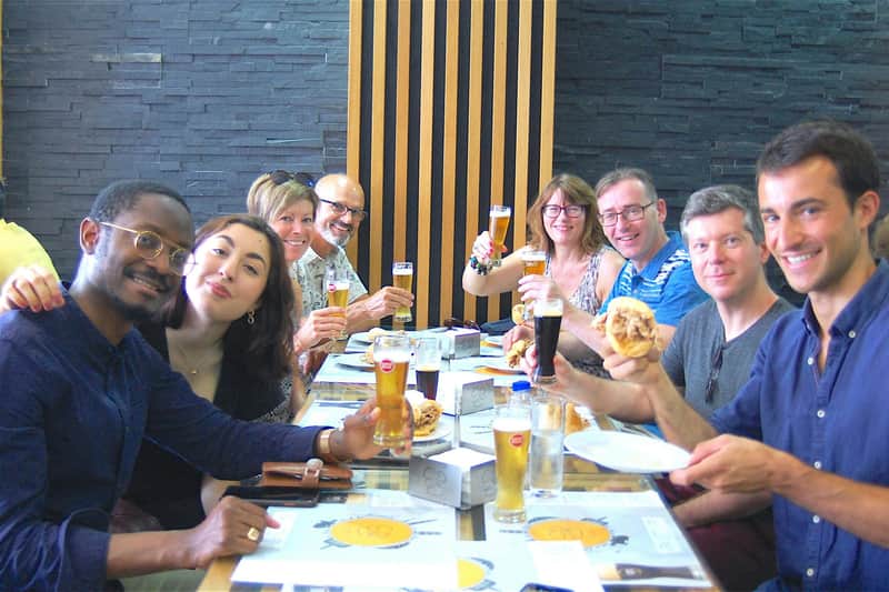 Porto: Guided Food and Wine Tour + Tasting A group of six people smiles and raises glasses of beer at a table, enjoying burgers in a restaurant with a modern decor.