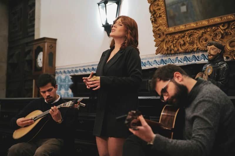 Porto: tour autoguidato del monastero + spettacolo di fado + drink (Porto) Una donna canta mentre due uomini suonano strumenti a corda in un ambiente interno con pareti e mobili decorativi.