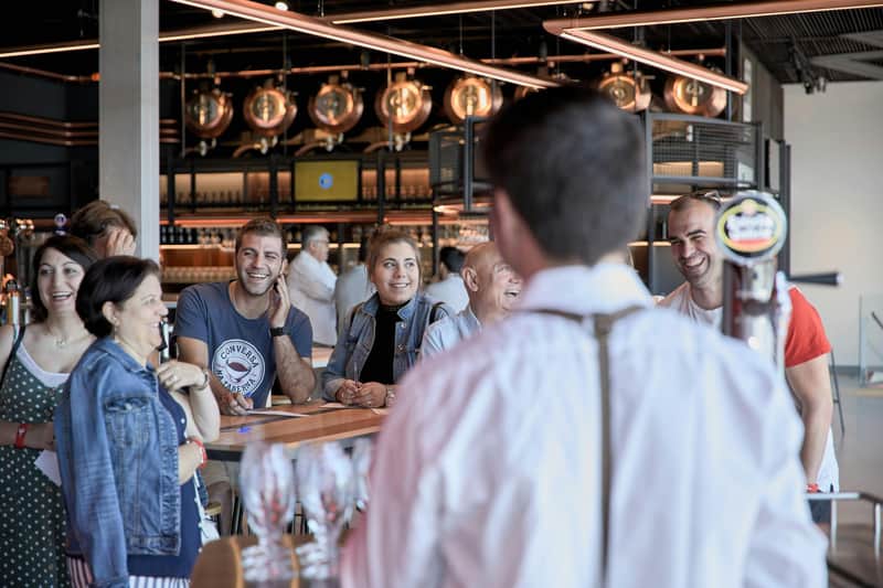 Un grupo de personas sonríe y conversa alrededor de una mesa de madera en un bar moderno con luces colgantes y cubas de cobre para la elaboración de cerveza.