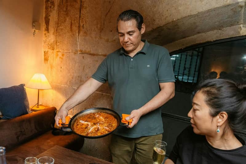 Barcelona: Gothic Quarter & El Born Guided Tapas Tour with Drinks A man serves a pan of food to a woman seated at a table in a rustic setting. The woman has a drink in front of her.