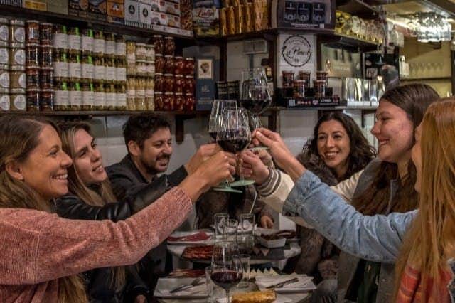 Barcelona: Guided Walking Tour + Tapas Tastings & Drinks A group of people seated at a table in a cozy restaurant, toasting with glasses of red wine, surrounded by shelves of food products.