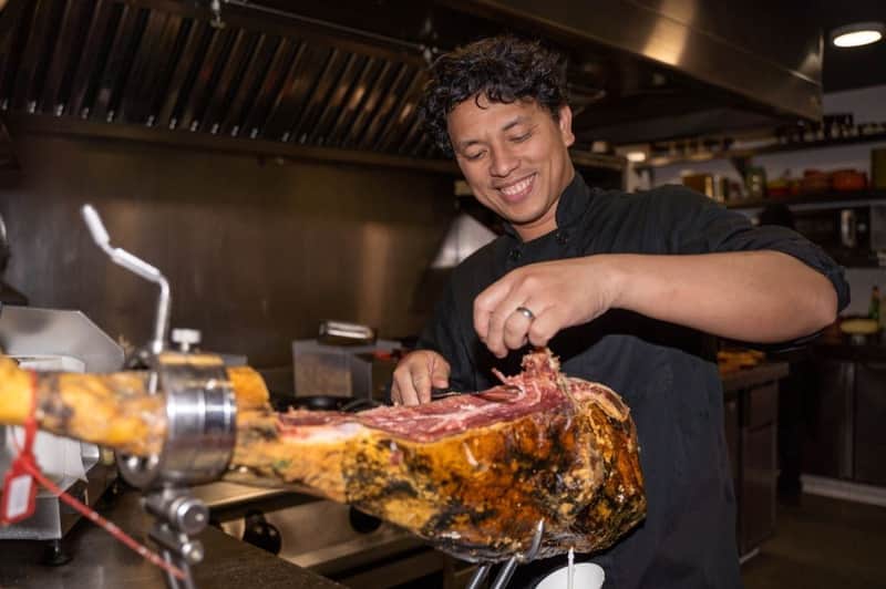 Barcelona: Small Group Guided Tapas & Wine Tour A chef smiles while slicing cured meat in a commercial kitchen.