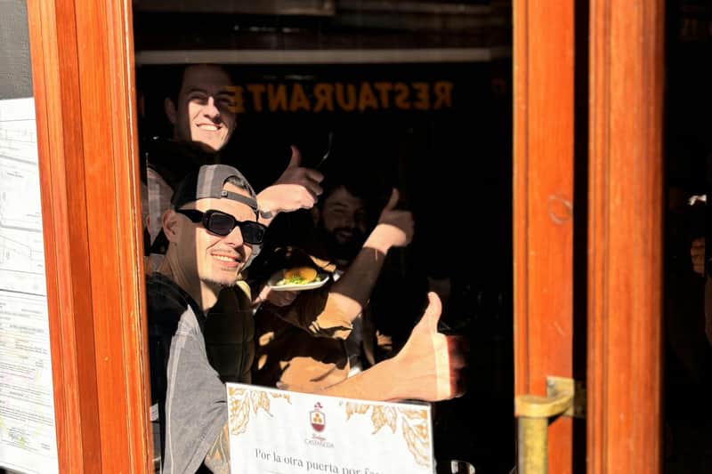 Granada: Guided Luxury Food Tour Three people behind a restaurant window giving thumbs-up, with one holding a plate. Menu and a sign visible on the window.