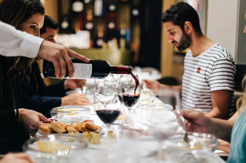 A person pours red wine into glasses at a dining table set with food, with people seated and dining in a softly lit room.