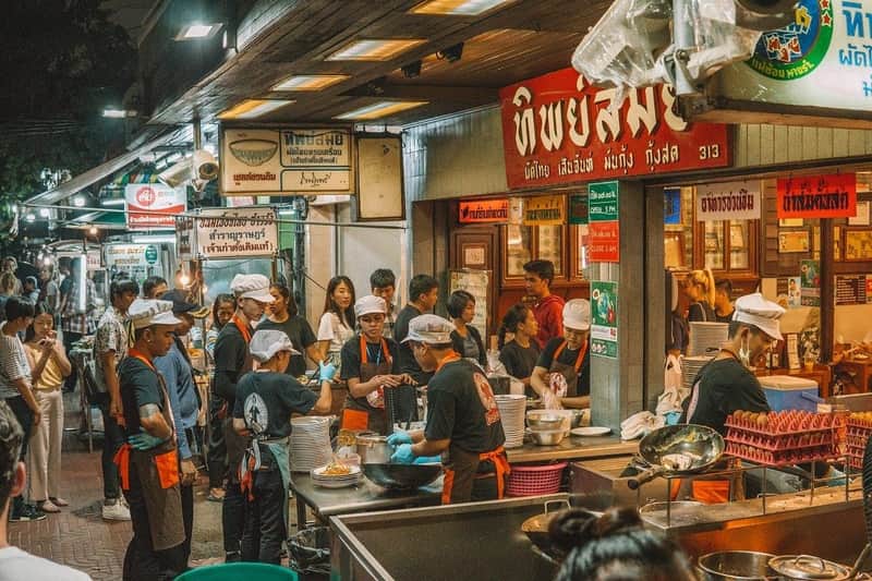 Bangkok by Night Tuk Tuk Adventure Busy street food stand with chefs in black shirts and white hats preparing meals, while customers wait in line. Signs in Thai above.