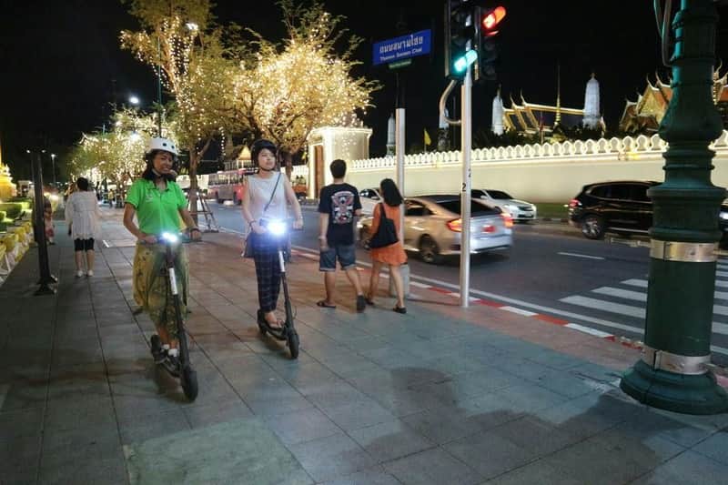 E-Scooter Night Tour: Electric Scooter Ride with Local Street Food Two people riding electric scooters on a lit street at night, with decorative lights on trees and pedestrians nearby.