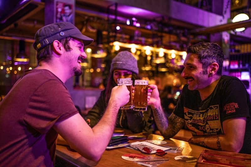 Three friends clink beer mugs in a lively, warmly lit pub, smiling and enjoying their time together.