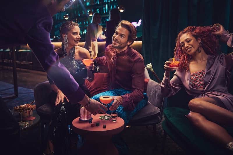 Three people enjoying cocktails in a dimly lit bar, sitting on armchairs, with a bartender and bar in the background.