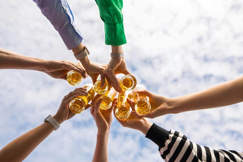 Several hands holding glasses filled with an amber-coloured liquid, raised together against a background of a cloudy sky.