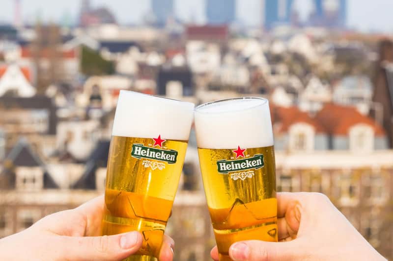 Two glasses of Heineken beer being clinked together, with a blurred urban cityscape in the background.