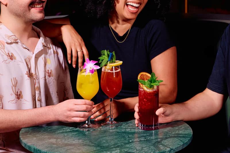 Three people enjoying colorful cocktails garnished with flowers, fruit, and mint at a green marble table.