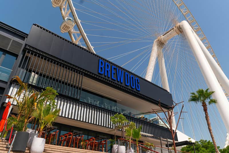 A modern BrewDog establishment with outdoor seating and large signage, located near a Ferris wheel under a clear sky.