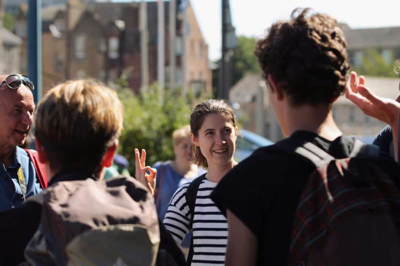 A group of people outdoors, one smiling person in a striped shirt gesturing while others listen, with buildings and greenery in the background.