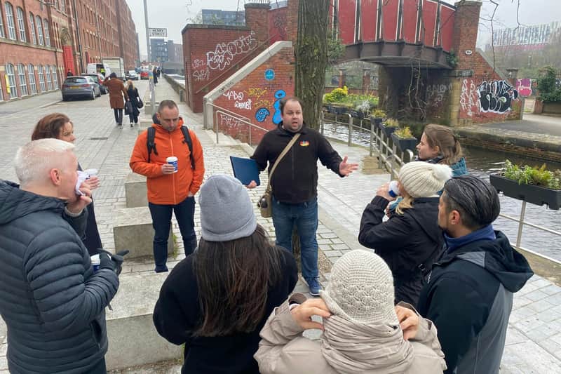 Un homme s'adresse à un petit groupe de personnes dans une rue couverte de graffitis, près d'un canal, avec un pont rouge en arrière-plan.