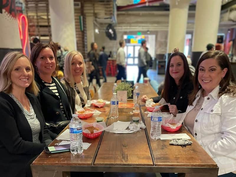 Four women seated at a wooden table in a casual dining environment, with food trays, water bottles, and decor on the table.