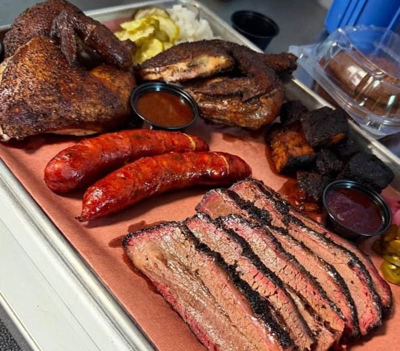 A platter of barbecue meats including brisket, sausage, chicken, and burnt ends, served with pickles and sauce cups.