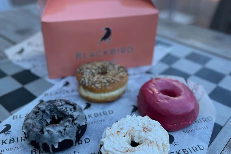 Vier verschiedene Donuts auf Blackbird-Papier vor einer rosa Schachtel mit dem Blackbird-Logo.