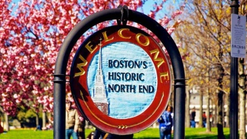 Ein rundes Schild mit der Aufschrift "Welcome Boston's Historic North End" und dem Bild eines Kirchturms vor dem Hintergrund des Frühlingslaubs.