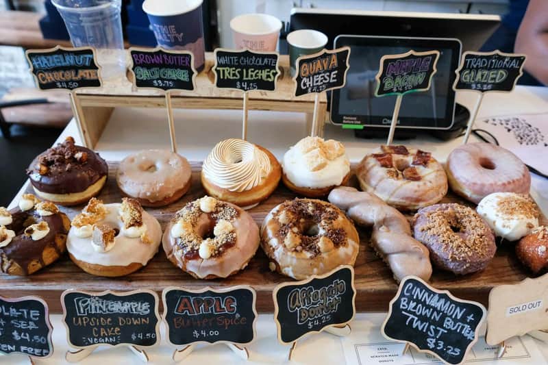 An assortment of gourmet donuts displayed with labels indicating different flavors such as brown butter, white chocolate tres leches, and maple bacon.