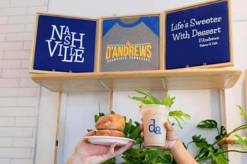 Two hands holding donuts on a plate and an iced drink in front of a shelf with three signs displaying Nashville-related text.