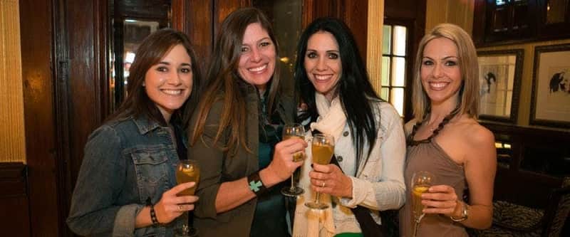 Two women smiling and holding glasses with beverages, standing in a warmly lit setting with wooden panels in the background.