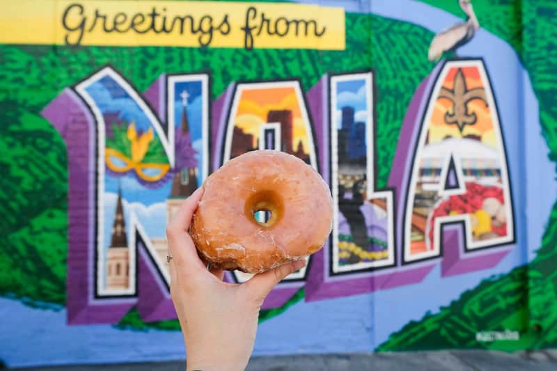 A hand holding a glazed doughnut in front of a colorful mural that reads "Greetings from NOLA."