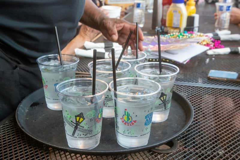 A tray holding six plastic cups of water with black straws, on a table with condiments, beads, and other items nearby.