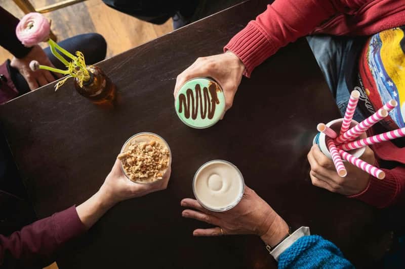 Three people holding different drinks and desserts at a wooden table, with only their hands visible.