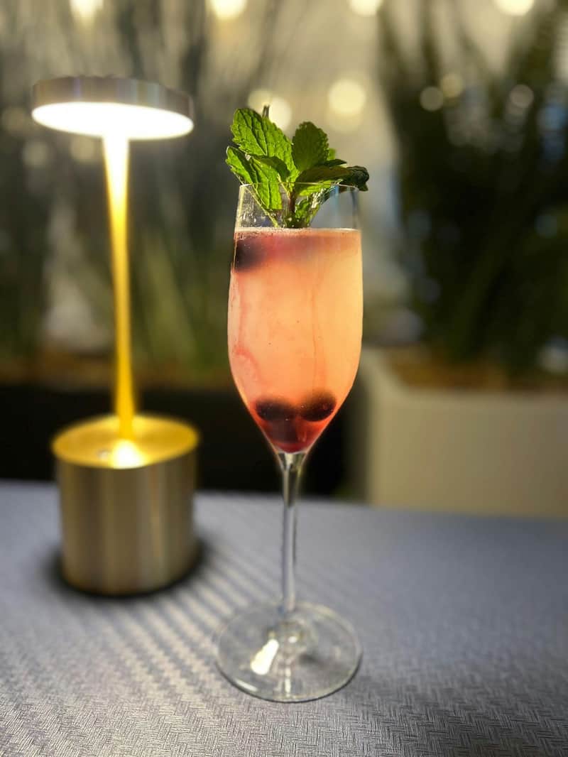 Craft Cocktail Gourmet Food Tour A pink cocktail in a flute glass garnished with mint leaves and berries, placed on a table near a warm-lit lamp.