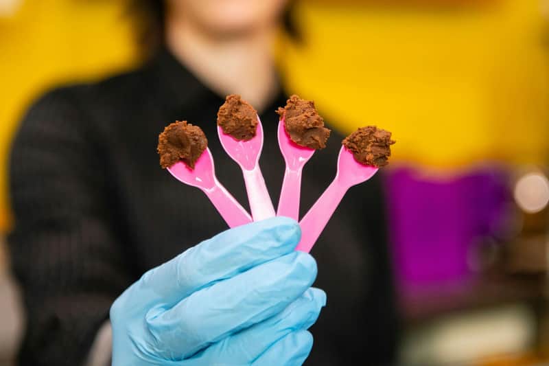 Quattro cucchiaini rosa con piccole porzioni di dessert al cioccolato, tenuti da una mano guantata su uno sfondo colorato.
