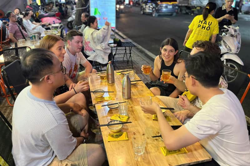 Ho Chi Minh City: Guided Craft Beer & Food Tour by Scooter A group of people sit around an outdoor wooden table with food and drinks, engaging in conversation at a busy street-side eatery.