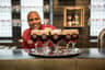 A smiling bartender in a red shirt stands behind a tray of six foamy dark beers on a counter.